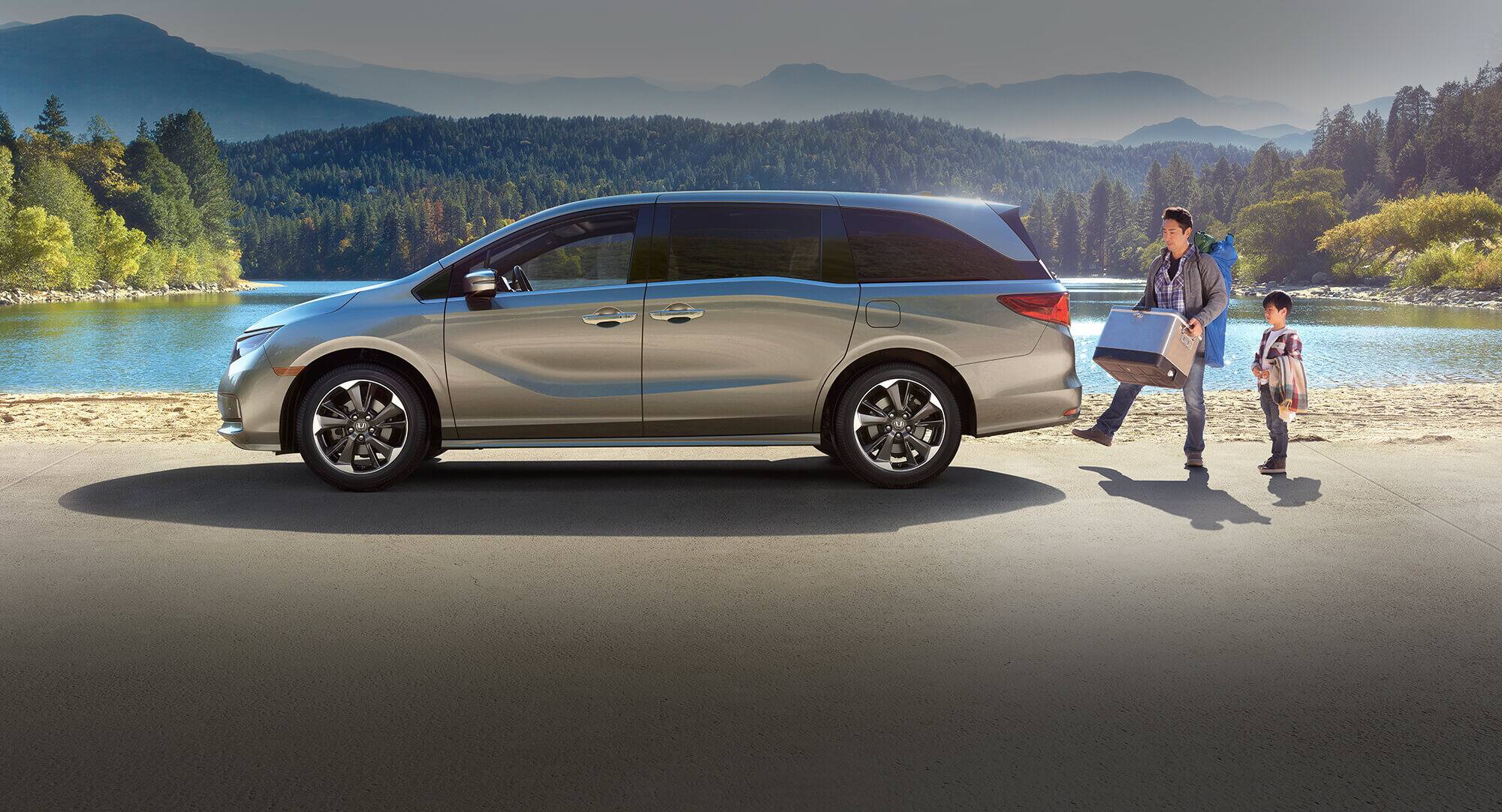 Driver-side profile view of the 2024 Honda Odyssey Elite in Lunar Silver Metallic parked in front of a lake, with a man activating the hands-free access power tailgate as a child looks on.